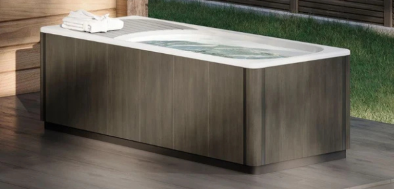 Minimalist outdoor cold plunge tub with grey cabinet and white shell, filled with water, placed on a wooden deck beside a modern building.